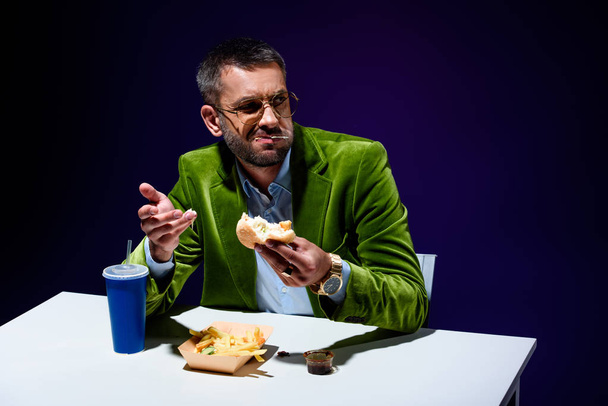 hombre en chaqueta de terciopelo comiendo hamburguesa en la mesa con gritos franceses y bebida de soda con fondo azul
 - Foto, Imagen