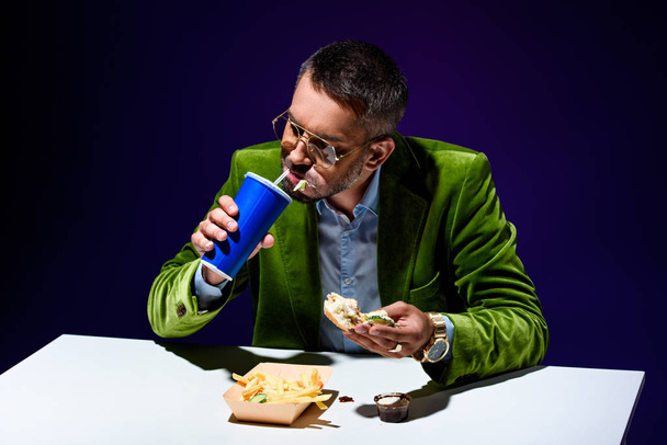 modischer Mann in Samtjacke trinkt Limonade, während er am Tisch mit Junk Food vor blauem Hintergrund sitzt - Foto, Bild
