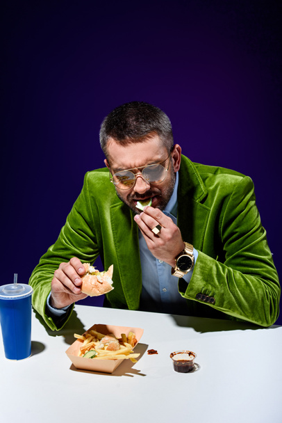 retrato de homem de veludo jaqueta comer hambúrguer à mesa com gritos franceses e refrigerante bebida com fundo azul
 - Foto, Imagem