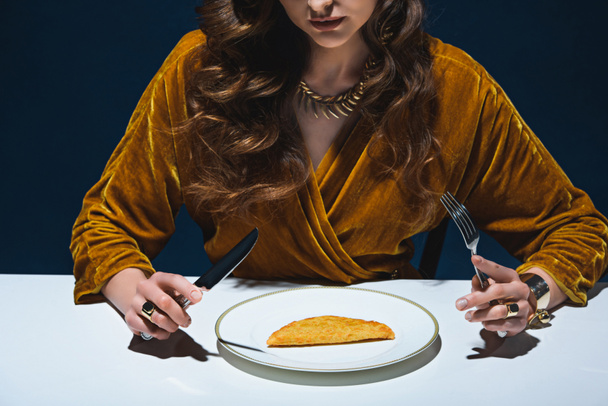 cropped shot of woman in luxury clothing sitting at table with meat pastry on plate with blue backdrop - Foto, Bild