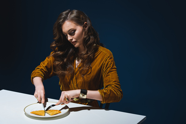 woman in stylish clothing cutting unhealthy cheburek at table with blue backdrop behind - Foto, afbeelding