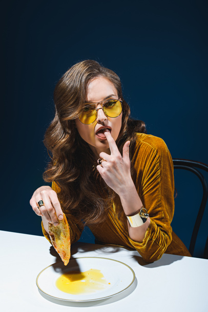 portrait of fashionable woman with unhealthy meat pastry sitting at table with blue backdrop - Photo, image
