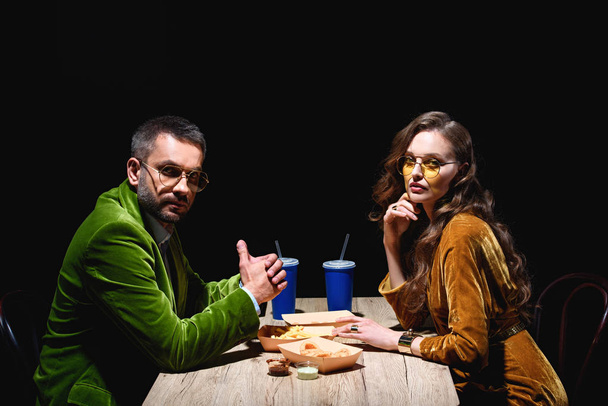 side view of couple in stylish velvet clothing sitting at table with fried onion rings, french fries and sauces with black background - Photo, Image