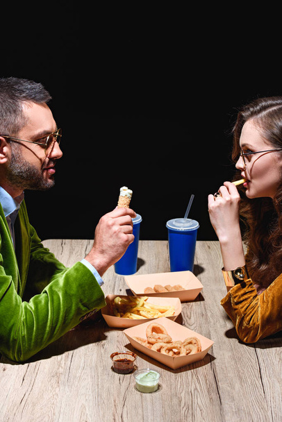 Seitenansicht eines Paares in stylischer Samtkleidung, das am Tisch mit schwarzem Hintergrund gebratene Zwiebelringe, Pommes frites und Soßen isst - Foto, Bild