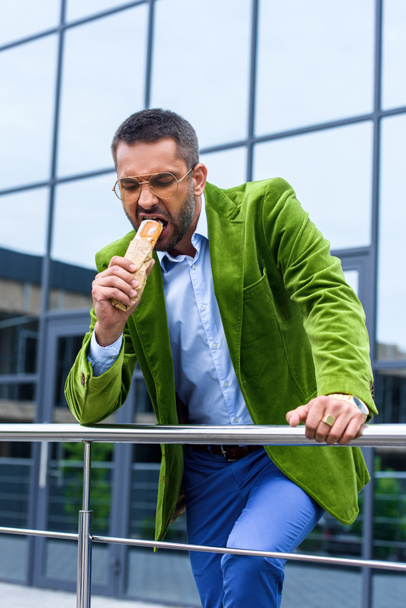 portrait of man in green velvet jacket eating french hot dog on street - Foto, afbeelding