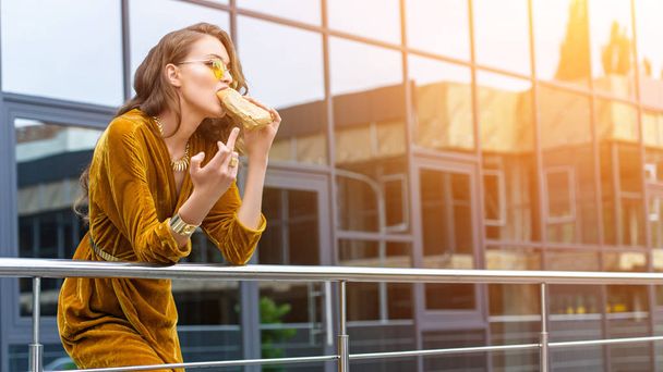 Frau in modischem Kleid isst französischen Hot Dog und zeigt Mittelfinger auf der Straße - Foto, Bild