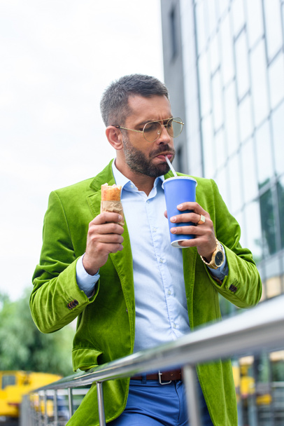 portrait of man in green velvet jacket with french hot dog drinking soda drink on street - Фото, изображение