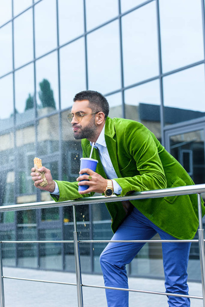 Вид сбоку человека в зеленой бархатной куртке с французским хот-догом и газированным напитком в руках на улице
 - Фото, изображение