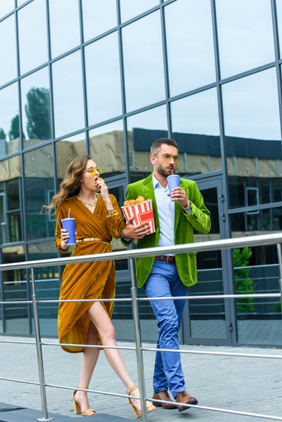 fashionable couple in velvet clothing with soda drinks eating fried chicken legs while walking on street - Fotografie, Obrázek