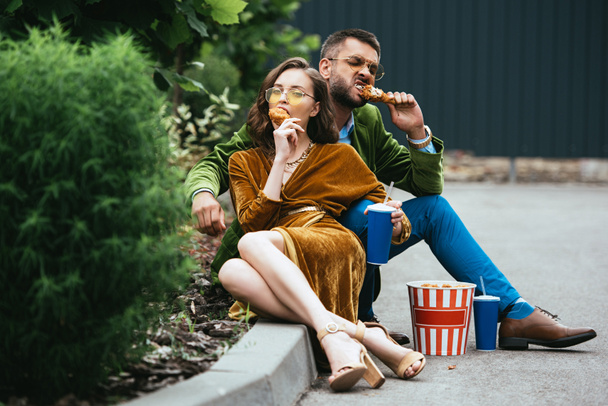fashionable couple in velvet clothing eating fried chicken legs on street - Photo, Image