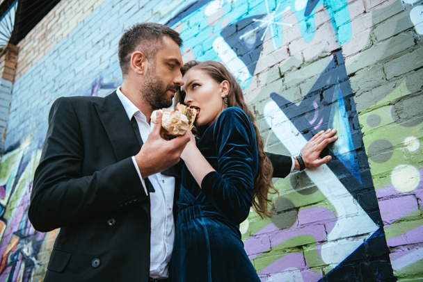 low angle view of couple in luxury clothing with hot dogs standing near graffiti on wall on street - Fotoğraf, Görsel