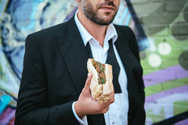 partial view of man in fashionable black jacket holding hot dog in hand on street - Foto, afbeelding