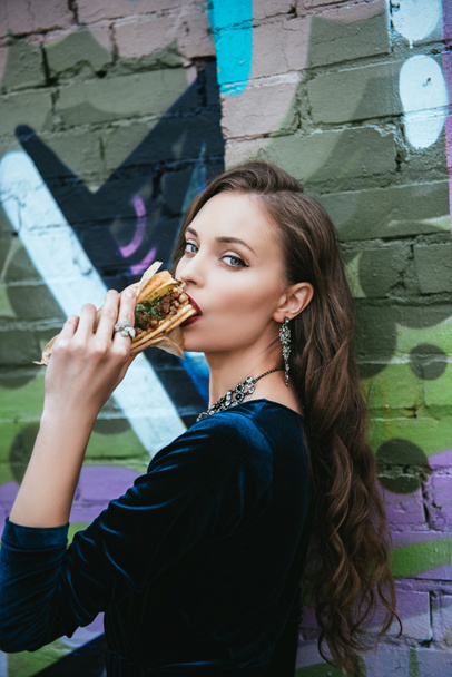 side view of beautiful woman in stylish clothing eating hot dog on street - Fotoğraf, Görsel
