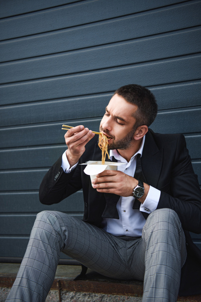 Porträt eines Mannes im schicken Anzug mit Essstäbchen, der auf der Straße asiatisches Essen isst - Foto, Bild
