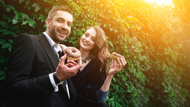 retrato de casal elegante sorridente com donuts de chocolate com folhagem verde atrás
 - Foto, Imagem