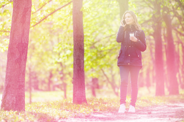 Talking on the phone, A girl is walking in the morning in a park with a smartphone and coffee, light toning - Photo, Image