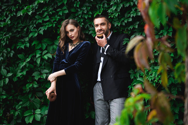 fashionable couple with chocolate doughnuts with green foliage behind looking at camera - Photo, image