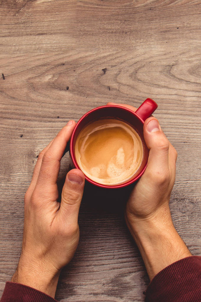 View from the top of two hands holding a red mug filled with coffee on a wooden table - Photo, Image