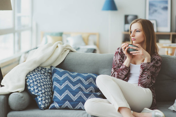 indoor lifestyle portrait of young woman relaxing at home with cup of hot tea or coffee, sitting on couch. Modern scandinavian apartment interior - Fotografie, Obrázek