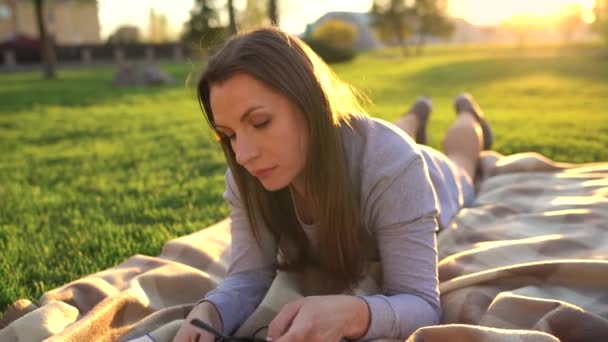 Girl in glasses reading book lying down on a blanket in the park at sunset - Filmati, video