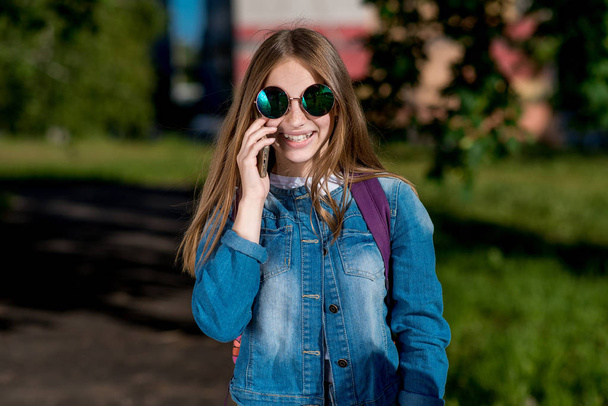 Küçük kız kız öğrenci genç. Doğa parkında yazın. Telefonda konuşuyorum. Kot elbise ve güneş gözlüğü. Duygu mutluluk sevinç ve gülümseme. - Fotoğraf, Görsel