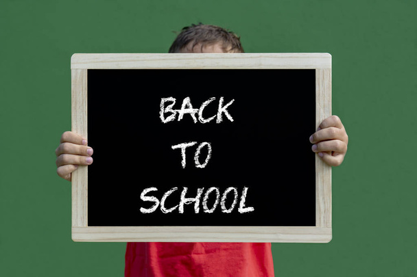 schoolchild holding small blackboard with text "back to school" against green background - Foto, immagini