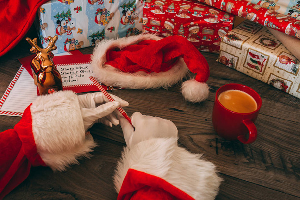 Kerstman met witte handschoenen holding een feestelijke potlood en een rode mok met koffie op zijn houten tafel met geschenken, inpakpapier, kerstmuts, en correspondentie. - Foto, afbeelding