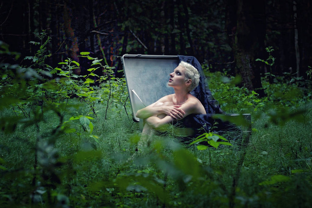A young girl with short white hair is sitting in a large suitcase in the middle of the forest - Foto, Imagem