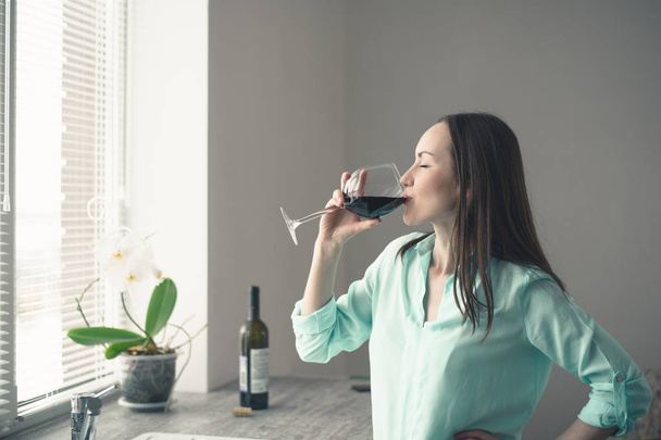 junges Mädchen steht am Fenster in der Küche und trinkt aus einem Glas Rotwein - Foto, Bild