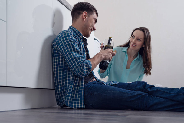 Fiatal pár ül a földön, a fehér konyha és ivott bort, szerelmes ünnepelni a housewarming. - Fotó, kép