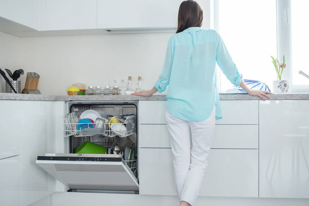 Жінка в білих джинсах і бірюзова сорочка стоїть з її спиною поруч з відкритою посудомийною машиною в інтер'єрі білої кухні перед вікном
. - Фото, зображення