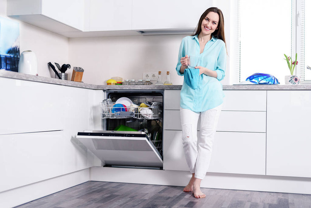 Full-length Portret van een gelukkige jonge huisvrouw in witte spijkerbroek en een turquoise shirt in een witte keuken staan met een handdoek in haar handen naast een open, moderne vaatwasser schoonmaken, afwassen, technologie. - Foto, afbeelding