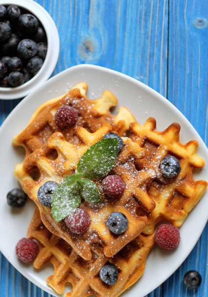 Traditional belgian waffles with fresh berrys and sugar powder on white plate, blue wooden background. Flat lay, top view, copy space. - Fotografie, Obrázek