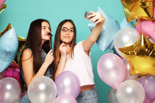Two teenage girls friends with colorfoul balloons make selfie on a phone. Blue background. - Valokuva, kuva