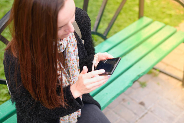 Nuori brunette istuu penkillä puistossa ja käyttää älypuhelinta. top view, käsite Internet, sosiaaliset verkostot, julkinen Wi-Fi
. - Valokuva, kuva