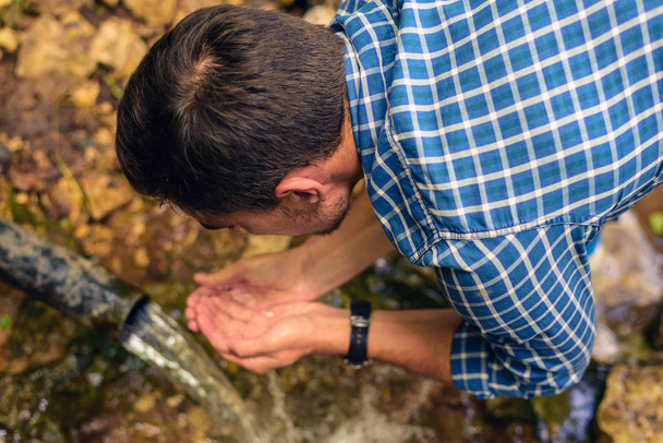 El hombre en la camisa en la jaula recoge el agua fresca del manantial en las manos dobladas, bebe el agua de la fuente
 - Foto, Imagen