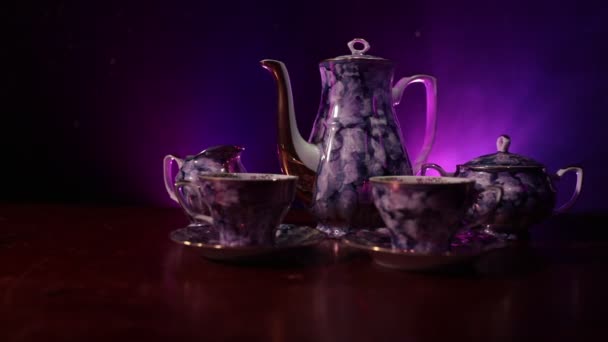 Coffee or tea ceremony conceptual theme. Old vintage ceramic tea or coffee pot with cups jug and sugar cup on dark toned background with light and smoke. Empty space for text. Slider shot - Footage, Video