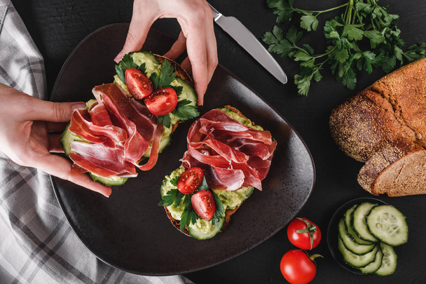 Woman's hands take a sandwich with prosciutto, avocado, cucumber, tomatoes on black stone table, decorated with napkin, knife. Clean eating, healthy breakfast. Top view, flat lay - Photo, image