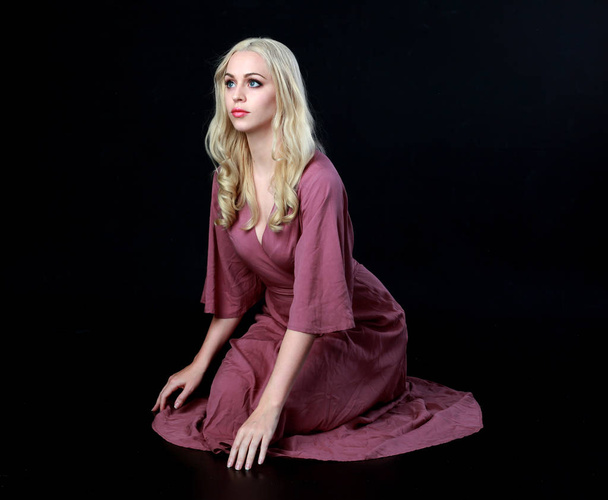 full length portrait of blonde girl wearing long purple dress. seated pose. black studio background. - Photo, Image