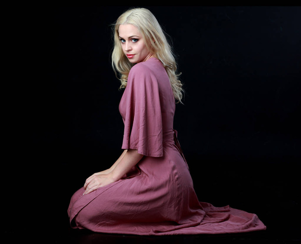 Ganzkörperporträt eines blonden Mädchens in einem langen lila Kleid. Sitzende Pose. schwarzer Studiohintergrund. - Foto, Bild