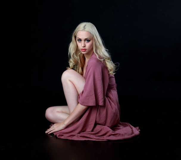Ganzkörperporträt eines blonden Mädchens in einem langen lila Kleid. Sitzende Pose. schwarzer Studiohintergrund. - Foto, Bild