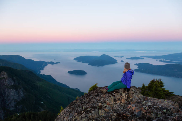 Приключенческая женщина на вершине горной скалы наслаждается прекрасным летним восходом солнца. Снято на горе Брансуик, залив Лайонс, к северу от Ванкувера, Британская Колумбия, Канада
. - Фото, изображение