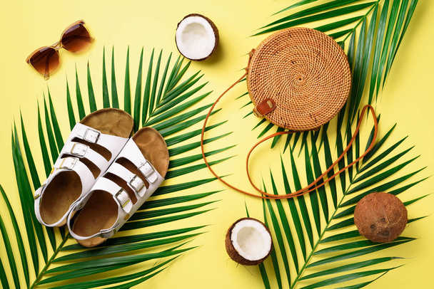 Round rattan bag, coconut, birkenstocks, palm branches, sunglasses on yellow background. Banner. Top view with copy space. Trendy bamboo bag and shoes. Summer fashion flat lay. Trip, vacation concept - Фото, изображение