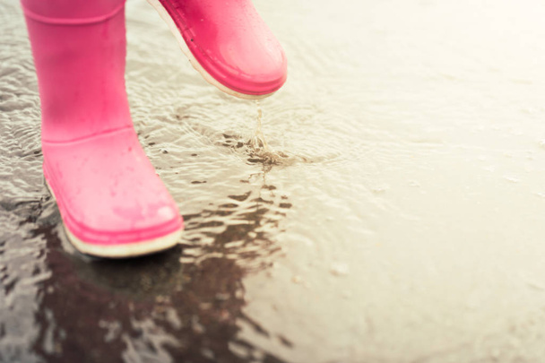 Feet of child in pink rubber boots jumping and splashing over puddle after rain. - Foto, Bild