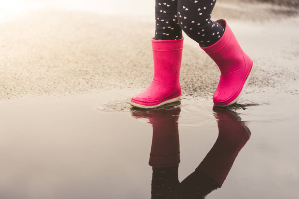 Kinderfüße in rosa Gummistiefeln springen und planschen nach Regen über Pfütze. - Foto, Bild