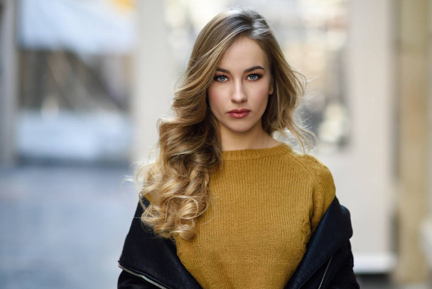 Blonde woman in urban background. Beautiful young girl wearing black leather jacket and mini skirt standing in the street. Pretty russian female with long wavy hair hairstyle and blue eyes. - Photo, Image