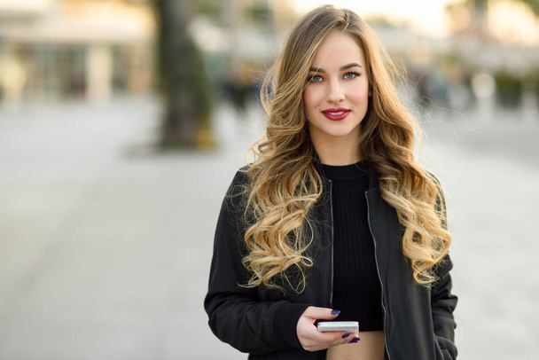 Blonde woman texting with her smart phone in urban background. Beautiful young girl wearing black jacket walking in the street. Pretty russian female with long wavy hair hairstyle. - Photo, Image