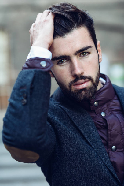 Young bearded man, model of fashion, in urban background wearing british elegant suit. Guy with beard and modern hairstyle touching his hair - 写真・画像