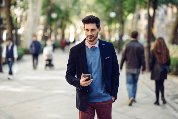 Hombre atractivo en la calle vistiendo traje elegante británico con teléfono inteligente en la mano. Joven hombre de negocios barbudo con peinado moderno en el fondo urbano
. - Foto, Imagen
