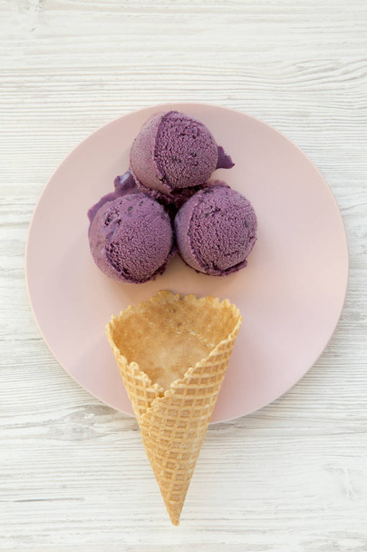 Cono de helado dulce con cucharadas de helado en un plato rosa sobre una superficie de madera blanca, vista superior. Desde arriba
. - Foto, imagen